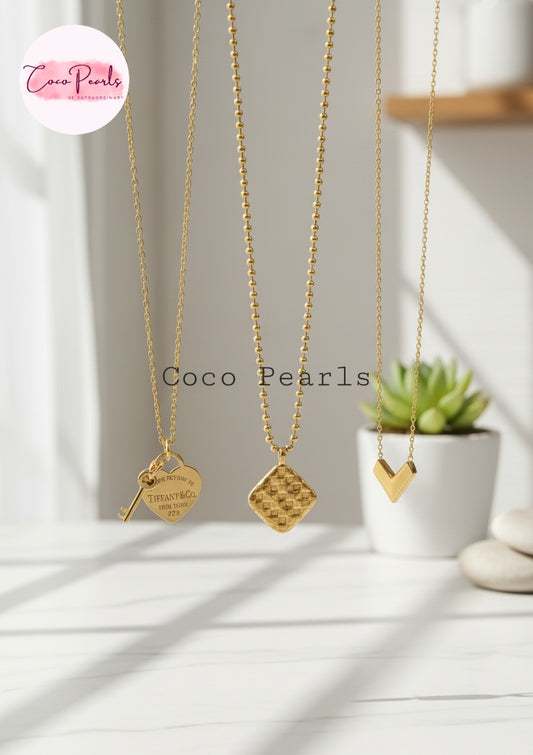 Coco Pearls Stainless Steel Anti Tarnish Three gold necklaces with different pendants on a white surface with a plant in the background