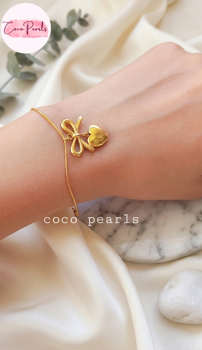 Stainless Steel anti tarnish Gold bracelet with a heart and bow design on a wrist, branded 'Coco Pearls', against a marble background.
