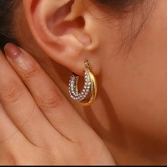 Coco pearls stainless Steel anti close-up of a person's ear wearing a gold crescent hoop earring adorned with small clear rhinestones, with the person's hand resting near the face.