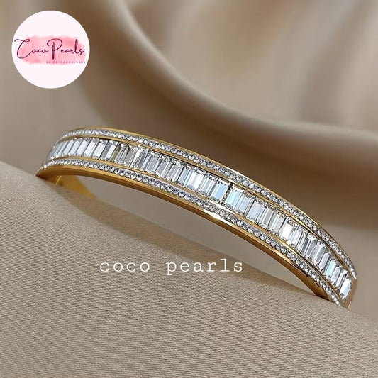 Coco pearls stainless Steel anti tarnish gold-plated eternity-style bangle bracelet with two rows of baguette-cut clear stones, displayed on a beige satin fabric background.