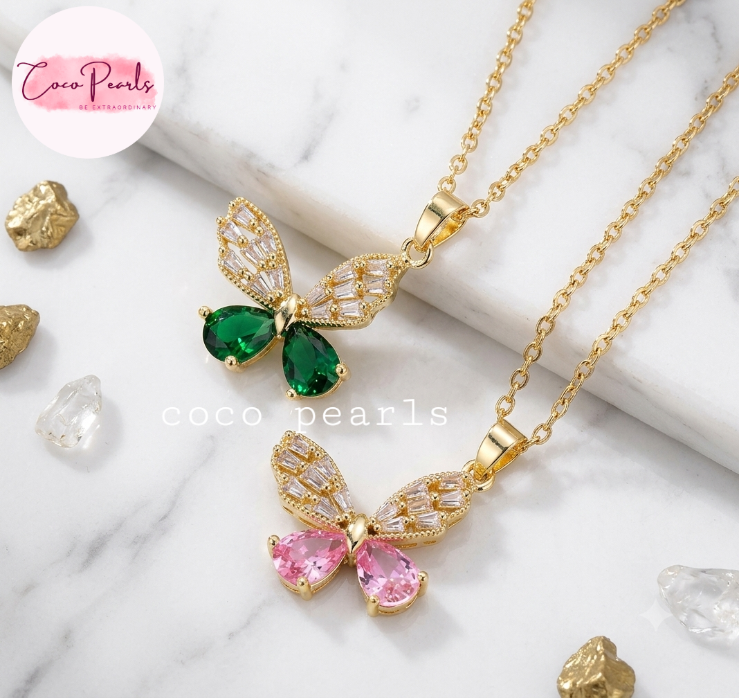 Coco pearls stainless Steel anti tarnish gold Two gold butterfly pendant necklaces on a white marble background, one with green teardrop gemstones and one with pink teardrop gemstones, with clear rhinestone wings.