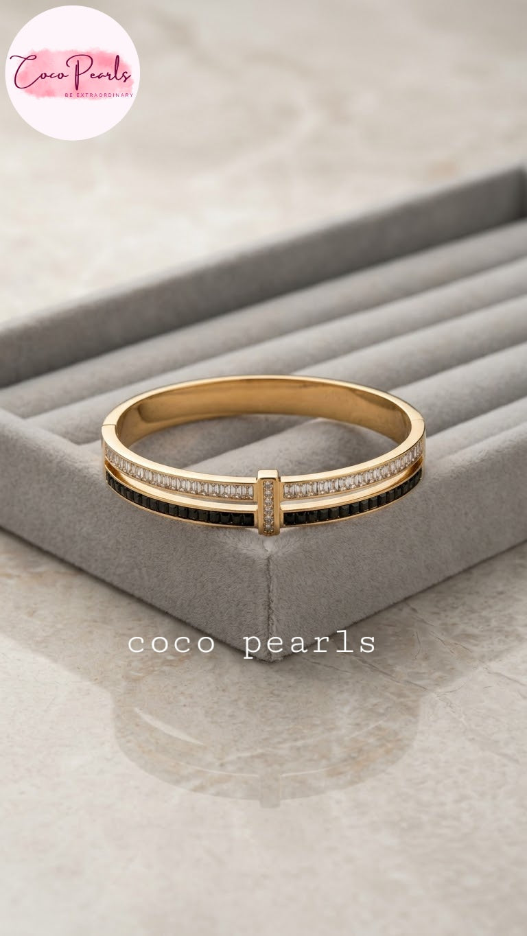 Coco pearls stainless Steel anti tarnish gold-plated bangle bracelet with a central black stripe and rows of clear stones on the front, displayed on a gray jewelry organizer tray.