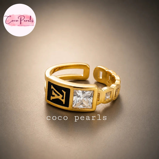 Coco pearls stainless Steel anti tarnish gold-plated ring with a black enamel panel featuring a gold LV monogram and a clear square gemstone, shown on a neutral brown background