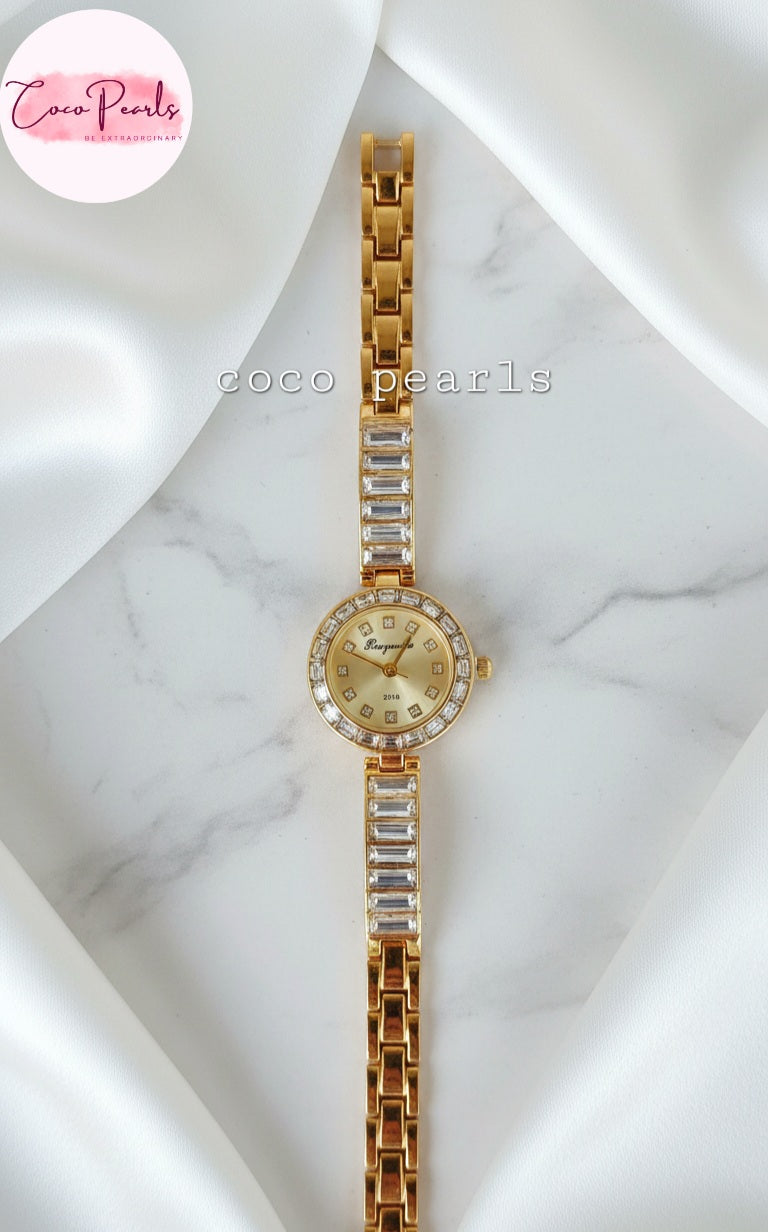 coco pearls Gold watch with rhinestone accents on a wrist against a marble background.