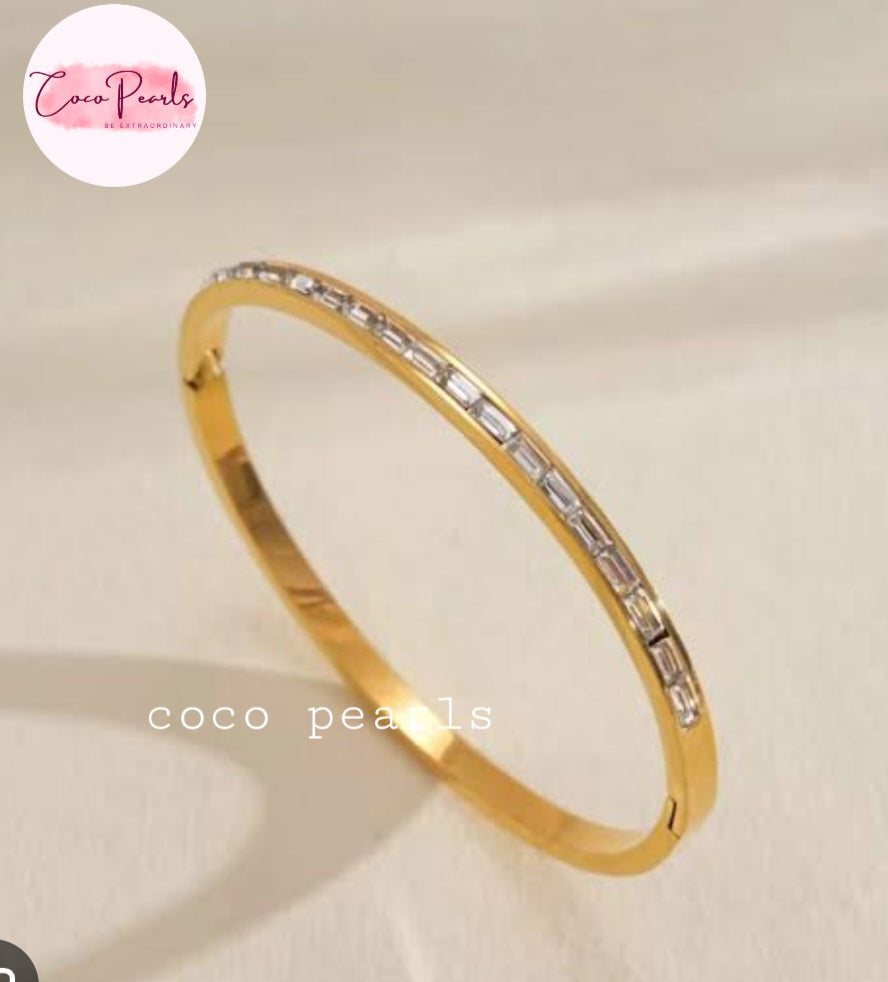 Coco pearls stainless Steel anti tarnish gold-plated bangle bracelet with a row of baguette-cut clear stones, displayed on a light beige fabric background. 