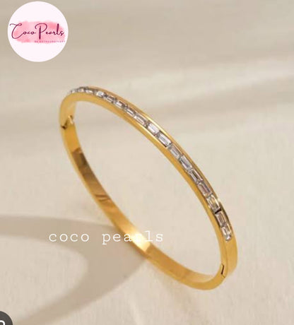 Coco pearls stainless Steel anti tarnish gold-plated bangle bracelet with a row of baguette-cut clear stones, displayed on a light beige fabric background. 