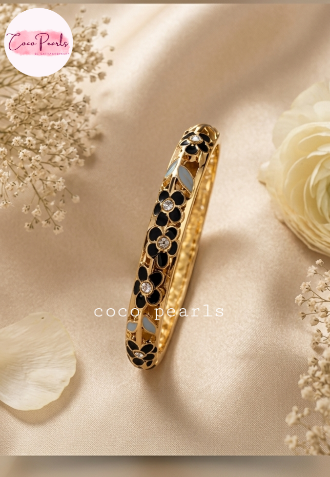 Coco pearls gold-plated bangle bracelet with black and white enamel floral detailing, displayed on a beige satin fabric with dried flowers