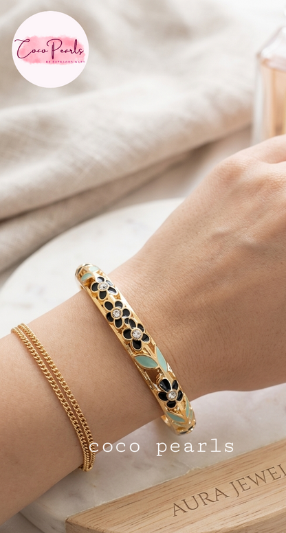 Coco pearls close-up of a person's wrist wearing a gold-plated bangle bracelet with black and white floral enameled details and light green leaf accents