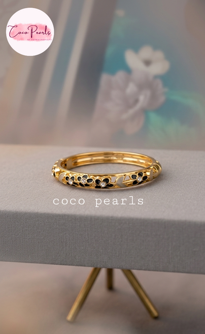 Coco pearls gold-plated bangle bracelet with black pearl-like inlays, displayed on a gray stand with a blurred floral background. 