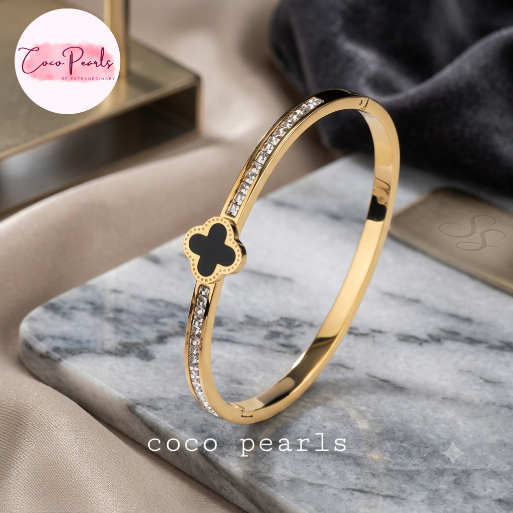 Coco pearls stainless Steel anti tarnish gold clover luxe diamond studded black clover charm in center handcuff displayed on marble surface 