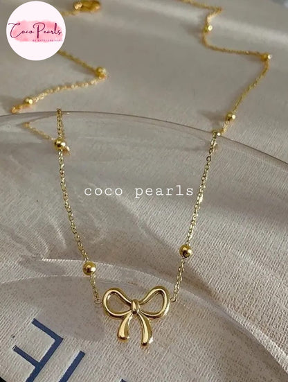 Coco pearls stainless Steel anti tarnish delicate gold necklace with a small bow pendant and small bead accents, displayed on a light beige textured fabric surface.