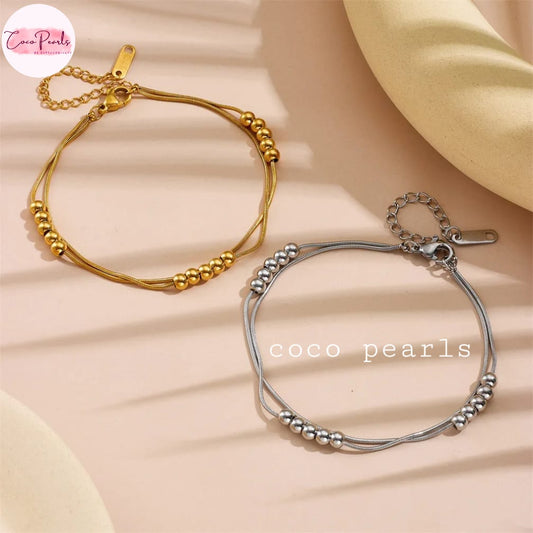 Coco pearls stainless Steel anti tarnish Two thin beaded bracelets, one in gold and one in silver, displayed on a soft beige background with a cream fabric prop.