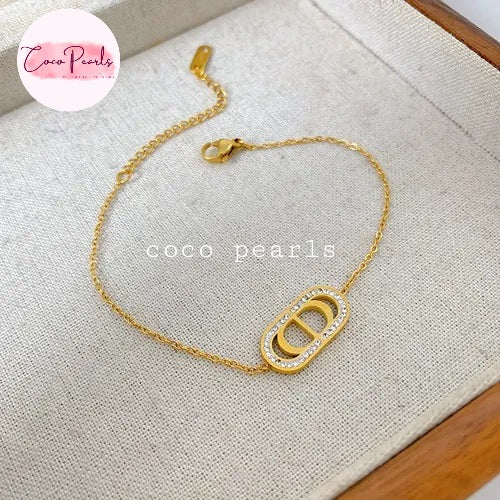 Coco pearls stainless Steel anti tarnish gold-tone chain bracelet with a large oval link centerpiece paved with small clear stones, displayed on a textured beige fabric surface.