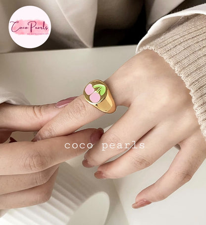 Coco pearls stainless Steel anti tarnish gold signet ring with a cherry design, worn on a person's ring finger. The ring features a polished gold band with a top plate decorated with two pink cherries on a green leaf.