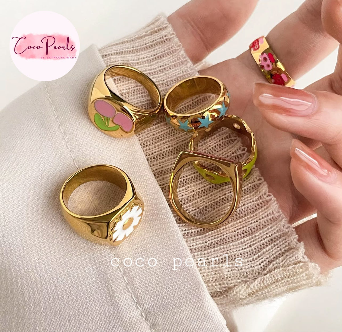 Coco pearls stainless Steel anti tarnish gold-plated signet rings with colorful enamel flower and leaf motifs are displayed on a hand resting on a beige knit sleeve.