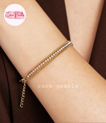 Coco pearls stainless Steel anti tarnish gold chain bracelet with a row of clear crystals, shown worn on a person's wrist against a dark brown background.