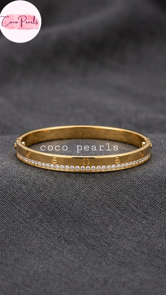 Coco pearls stainless Steel anti tarnish gold-plated bangle bracelet with a row of clear faux-pearls and engraved lettering, displayed on a dark gray textured fabric background.