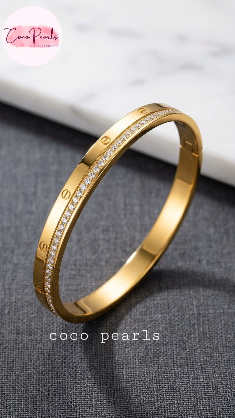 Coco pearls stainless Steel anti tarnish gold bangle bracelet with a row of small clear stones and screw-head details, displayed on a dark textured surface with a white marble background. 
