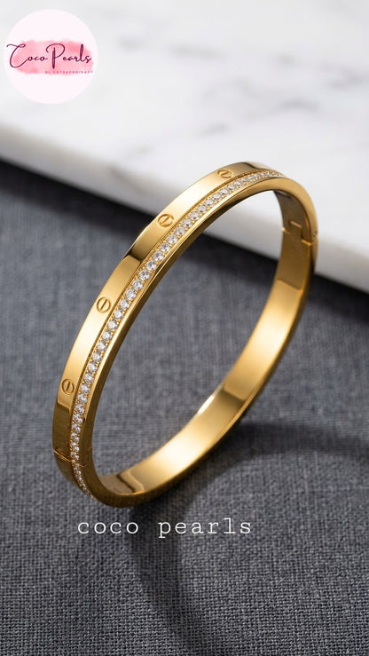 Coco pearls stainless Steel anti tarnish gold bangle bracelet with a row of small clear stones and screw-head details, displayed on a dark textured surface with a white marble background. 