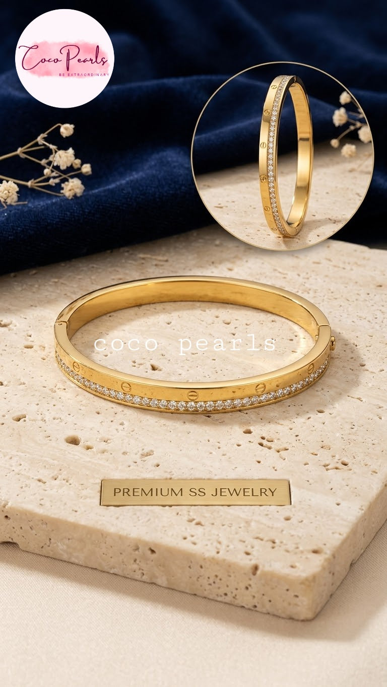 Coco pearls stainless Steel anti tarnish gold bangle bracelet with a row of small clear stones along the front, displayed on a beige stone slab.