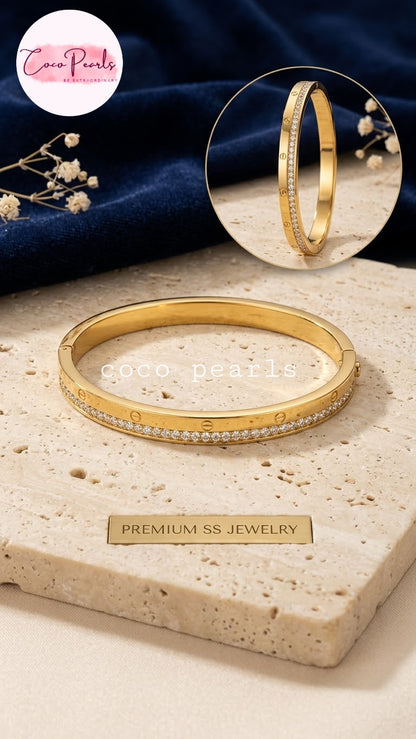 Coco pearls stainless Steel anti tarnish gold bangle bracelet with a row of small clear stones along the front, displayed on a beige stone slab.