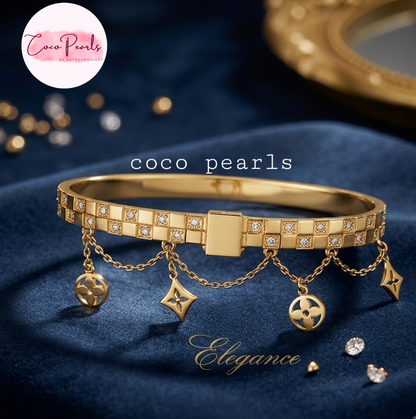 Coco pearls stainless Steel anti tarnish gold-tone Coco Pearls bangle bracelet with rhinestone-set segments and dangling chain charms, displayed on a dark blue satin fabric background with a gold mirror edge in the background.