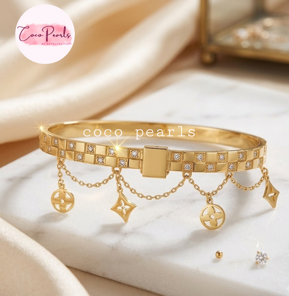 Coco pearls stainless Steel anti tarnish gold bangle bracelet with a geometric pattern of clear rhinestones, featuring three dangling chain charms with circular and star-shaped motifs. The bracelet is displayed on a white marble slab with draped beige satin fabric in the background.