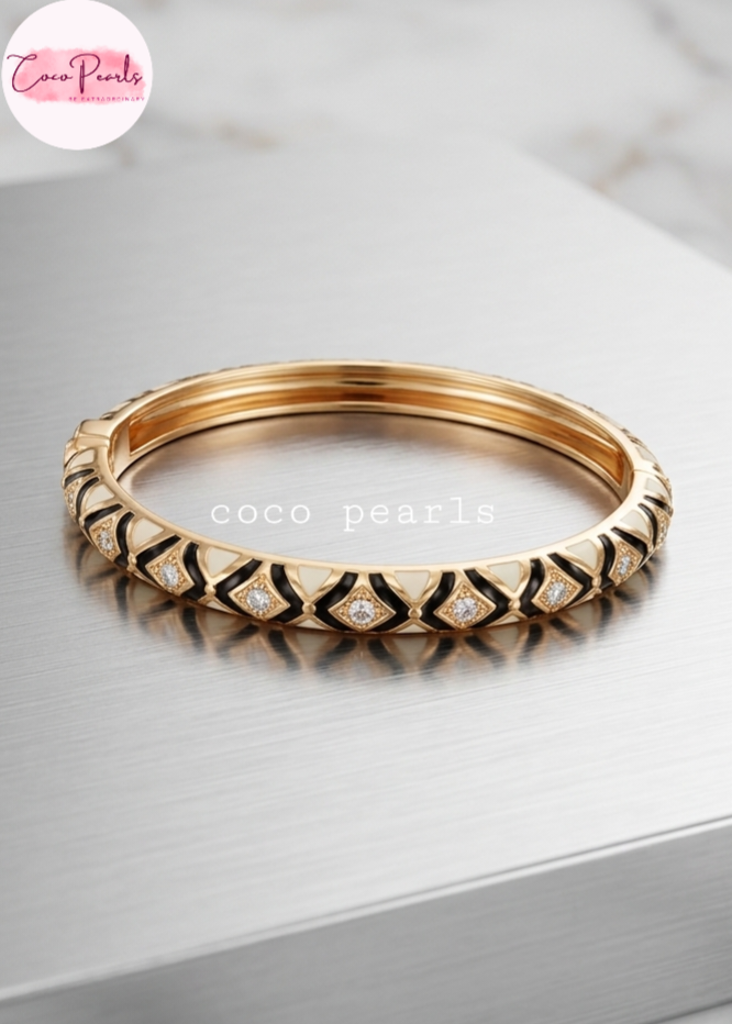 Coco pearls gold-plated bangle bracelet with black enamel geometric patterns and small clear pavé-set stones, displayed on a brushed silver metal surface.