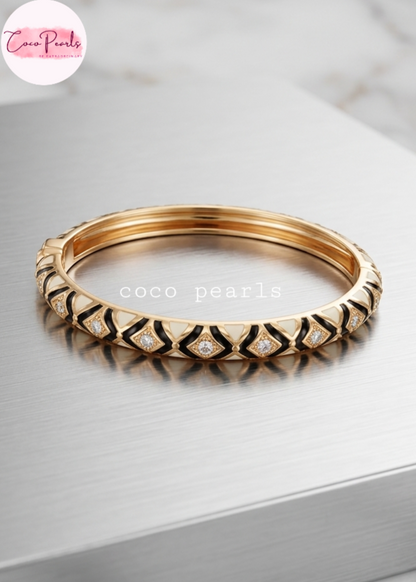 Coco pearls gold-plated bangle bracelet with black enamel geometric patterns and small clear pavé-set stones, displayed on a brushed silver metal surface.