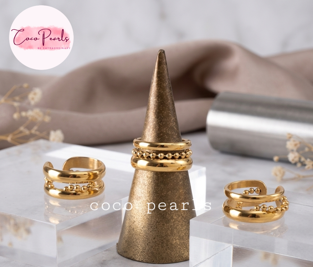 Coco pearls stainless Steel anti tarnish Three gold-toned open cuff rings displayed on clear acrylic blocks, with one ring centered on a metallic cone stand. 