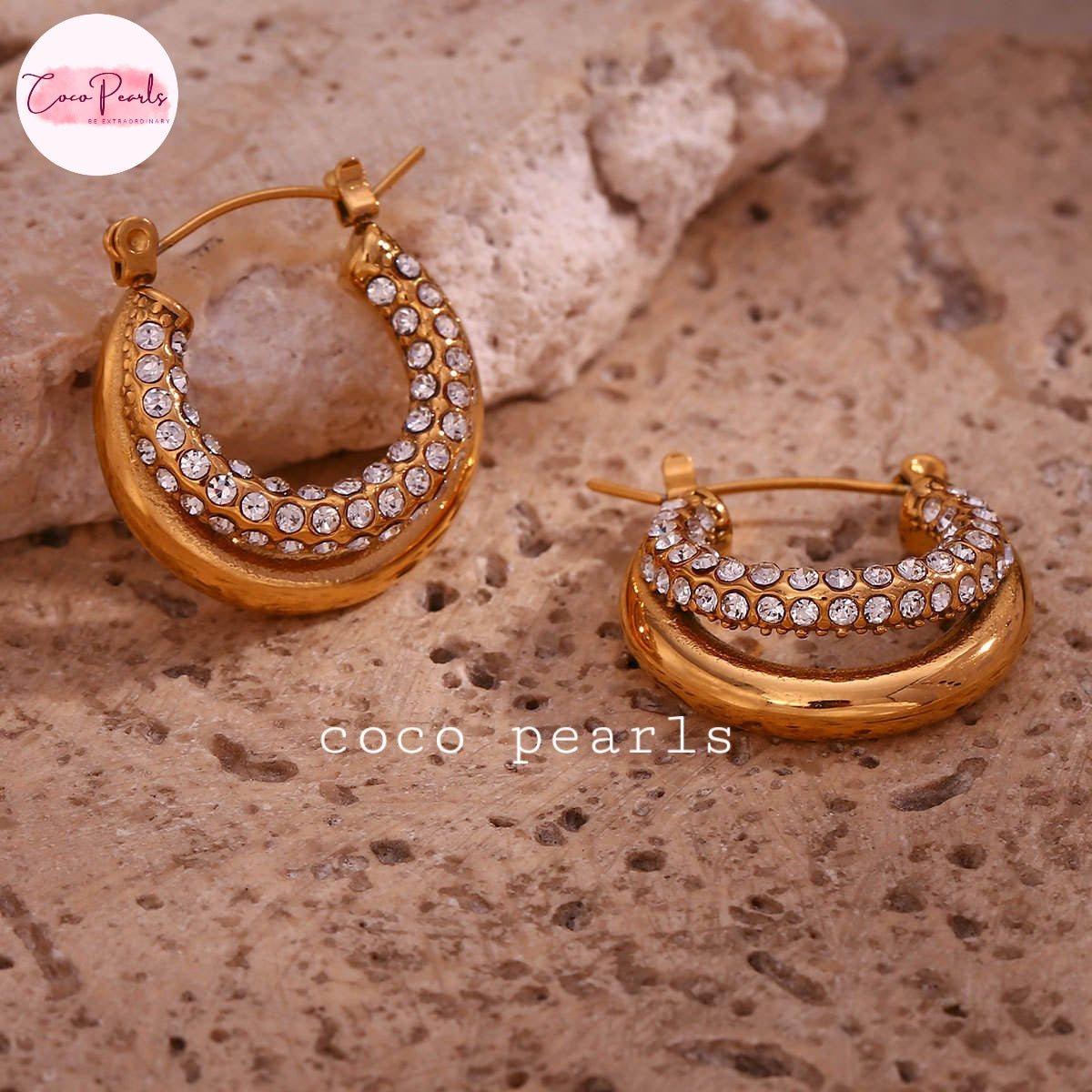 Coco pearls stainless Steel anti tarnish pair of gold crescent hoop earrings adorned with rows of clear rhinestones, displayed on a textured beige stone surface.