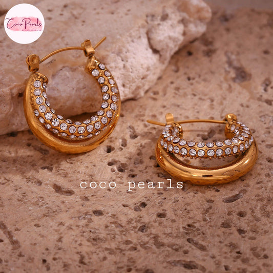 Coco pearls stainless Steel anti tarnish pair of gold crescent hoop earrings adorned with rows of clear rhinestones, displayed on a textured beige stone surface.