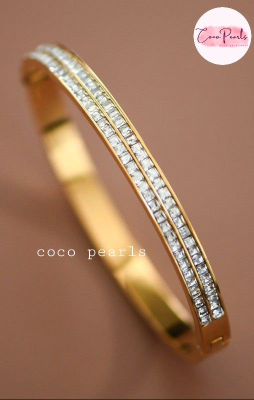 Coco pearls stainless Steel anti tarnish gold-plated hinged bangle bracelet with two parallel rows of clear pavé-set stones, shown on a warm brown background