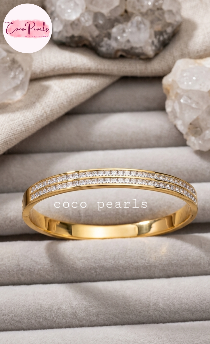 Coco pearls stainless Steel anti tarnish gold bangle bracelet with two rows of pavé-set clear stones, displayed on a gray jewelry tray with crystals and fabric in the background.