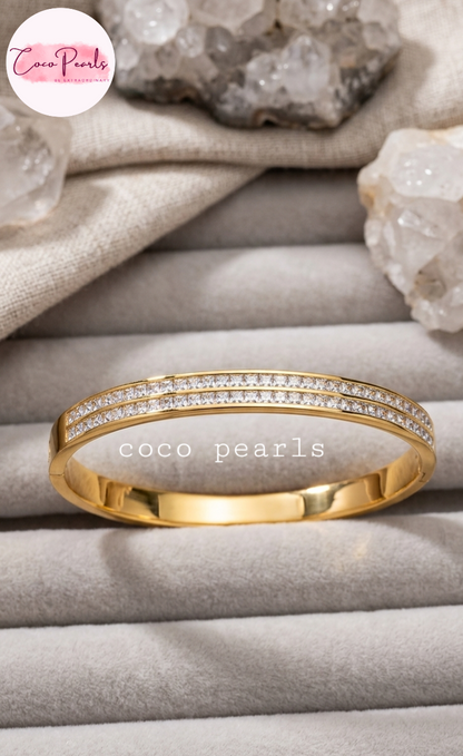 Coco pearls stainless Steel anti tarnish gold bangle bracelet with two rows of pavé-set clear stones, displayed on a gray jewelry tray with crystals and fabric in the background.