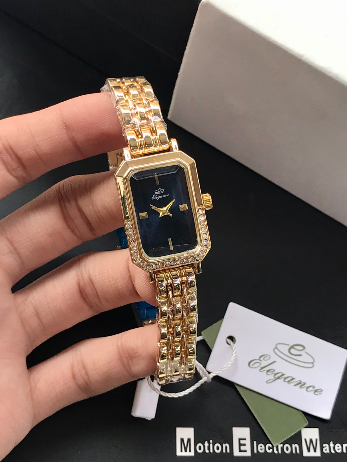 Coco pearls stainless Steel anti tarnish gold-tone women's watch with a rectangular bezel, crystal-embellished edges, and a black dial, held in a hand. The watch has a matching gold-tone link bracelet.