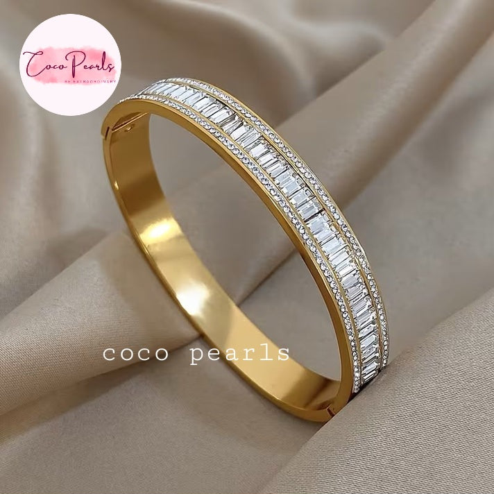 Coco pearls stainless Steel anti tarnish gold bangle bracelet with two rows of baguette-cut clear stones, displayed on draped beige satin fabric.