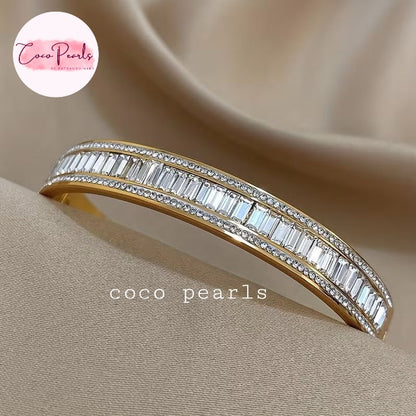 Coco pearls stainless Steel anti tarnish gold-plated eternity-style bangle bracelet with two rows of baguette-cut clear stones, displayed on a beige satin fabric background.