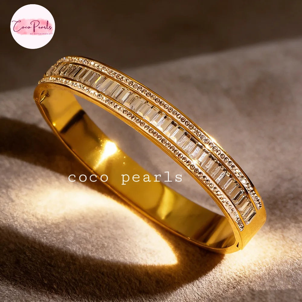 Coco pearls stainless Steel anti tarnish gold bangle bracelet with two rows of baguette-cut clear stones, photographed on a textured beige surface with dramatic warm lighting. 