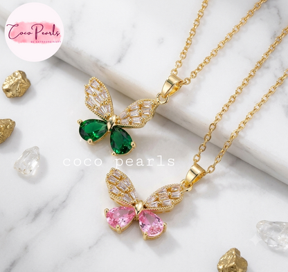 Coco pearls stainless Steel anti tarnish gold Two gold butterfly pendant necklaces on a white marble background, one with green teardrop gemstones and one with pink teardrop gemstones, with clear rhinestone wings.