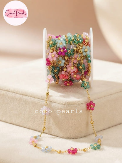 Coco pearls stainless Steel anti tarnish spool of beaded jewelry chain from Coco Pearls, featuring colorful pastel beads in pink, green, and blue with gold-toned links and flower charms, displayed on a beige jewelry stand.