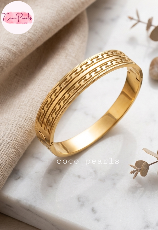 Coco pearls stainless Steel anti tarnish gold bangle bracelet with a detailed geometric pattern, displayed on a marble surface next to a beige linen cloth.