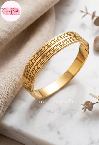 Coco pearls stainless Steel anti tarnish gold bangle bracelet with a detailed geometric pattern, displayed on a marble surface next to a beige linen cloth.