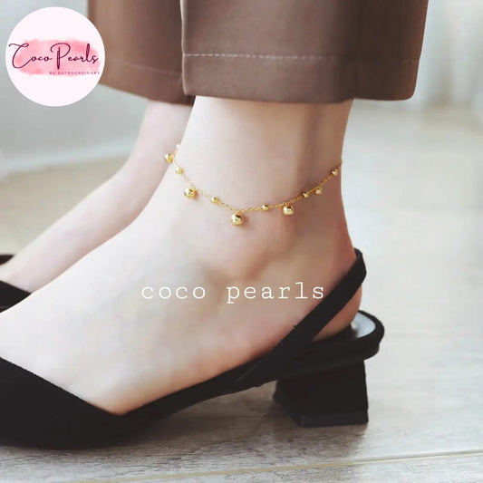 Coco pearls stainless Steel anti tarnish delicate gold chain anklet with small bead charms worn on a person's ankle, paired with black slingback mule sandals and brown trousers.