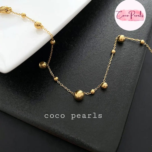 Coco pearls stainless Steel anti tarnish delicate gold chain necklace with small polished beads and one larger central bead, displayed on a black textured surface with a white geometric prop.