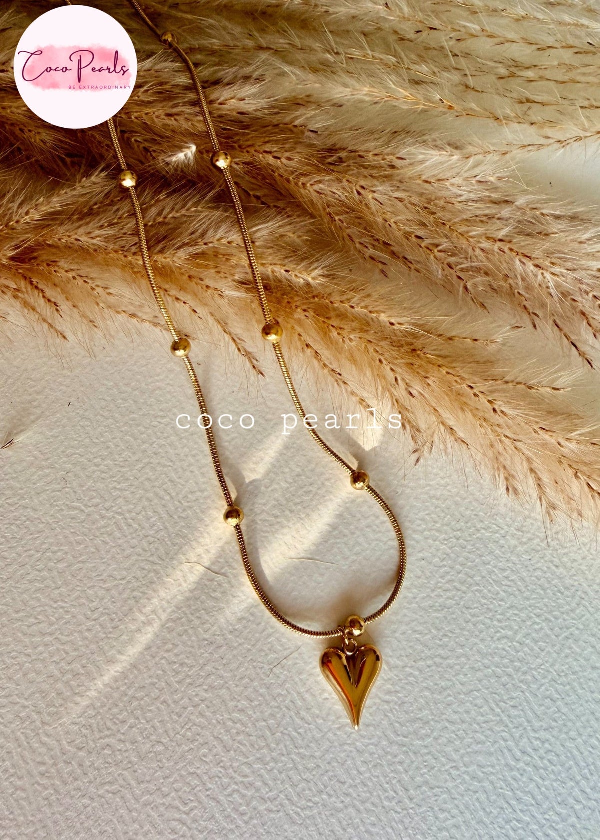 Coco pearls stainless Steel anti tarnish delicate gold necklace with a small heart pendant, laid flat on a textured white surface with dried pampas grass.
