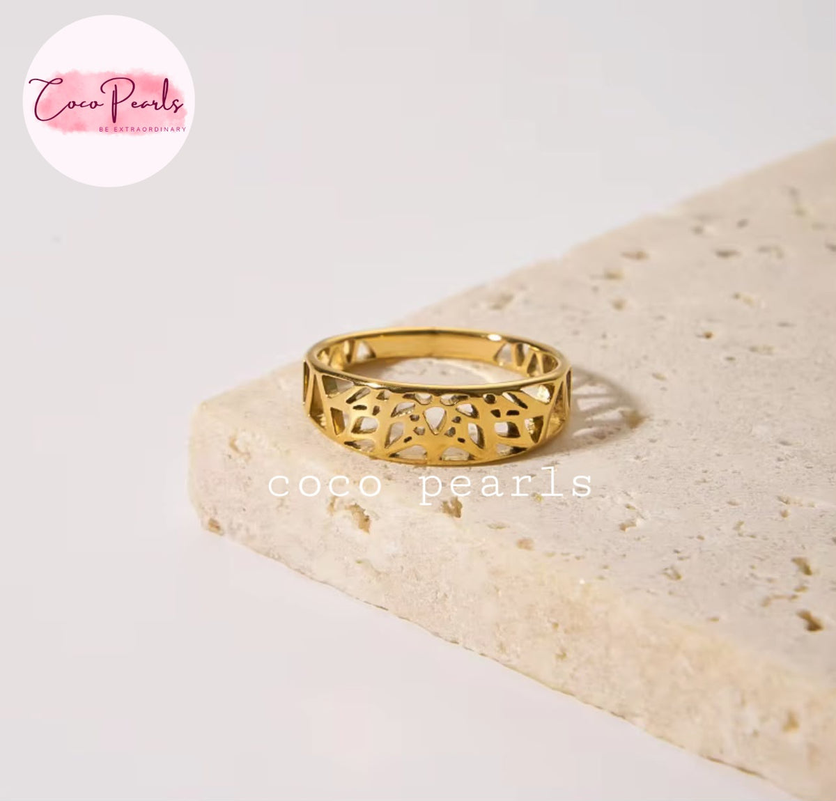 Coco pearls stainless Steel anti tarnish gold ring with an intricate openwork filigree pattern, resting on a textured beige stone slab against a clean white background.