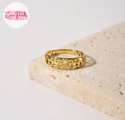 Coco pearls stainless Steel anti tarnish gold ring with an intricate openwork filigree pattern, resting on a textured beige stone slab against a clean white background.