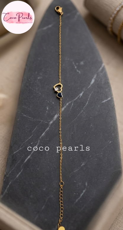 Coco pearls stainless Steel anti tarnish delicate gold chain bracelet with a small heart-shaped charm featuring a dark blue heart and a clear stone, displayed on a dark gray stone slab. 