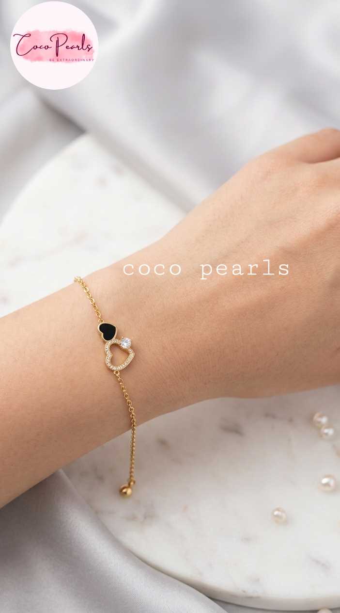Coco pearls stainless Steel anti tarnish gold chain bracelet with a black heart and a heart-shaped pearl charm, worn on a person's wrist. The background consists of a white marble tray and soft white fabric.
